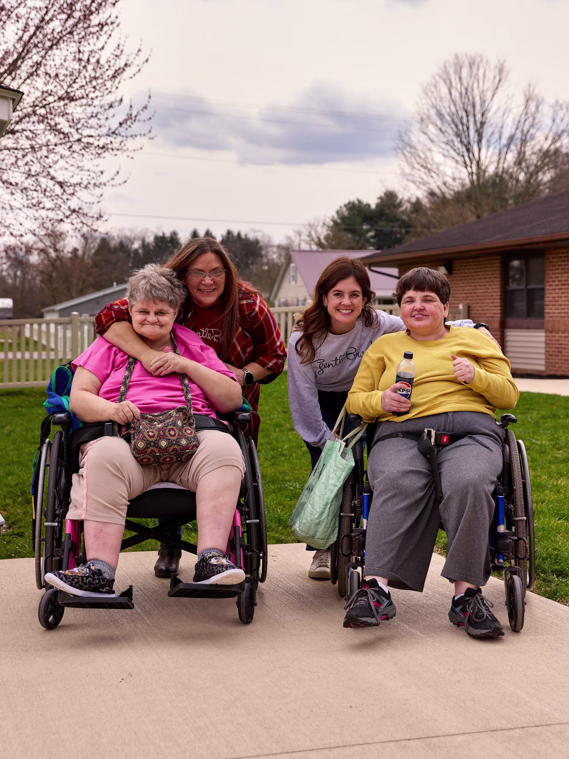 Two women in wheelchairs outdoors smiling, each accompanied by a standing woman hugging or leaning close behind them.