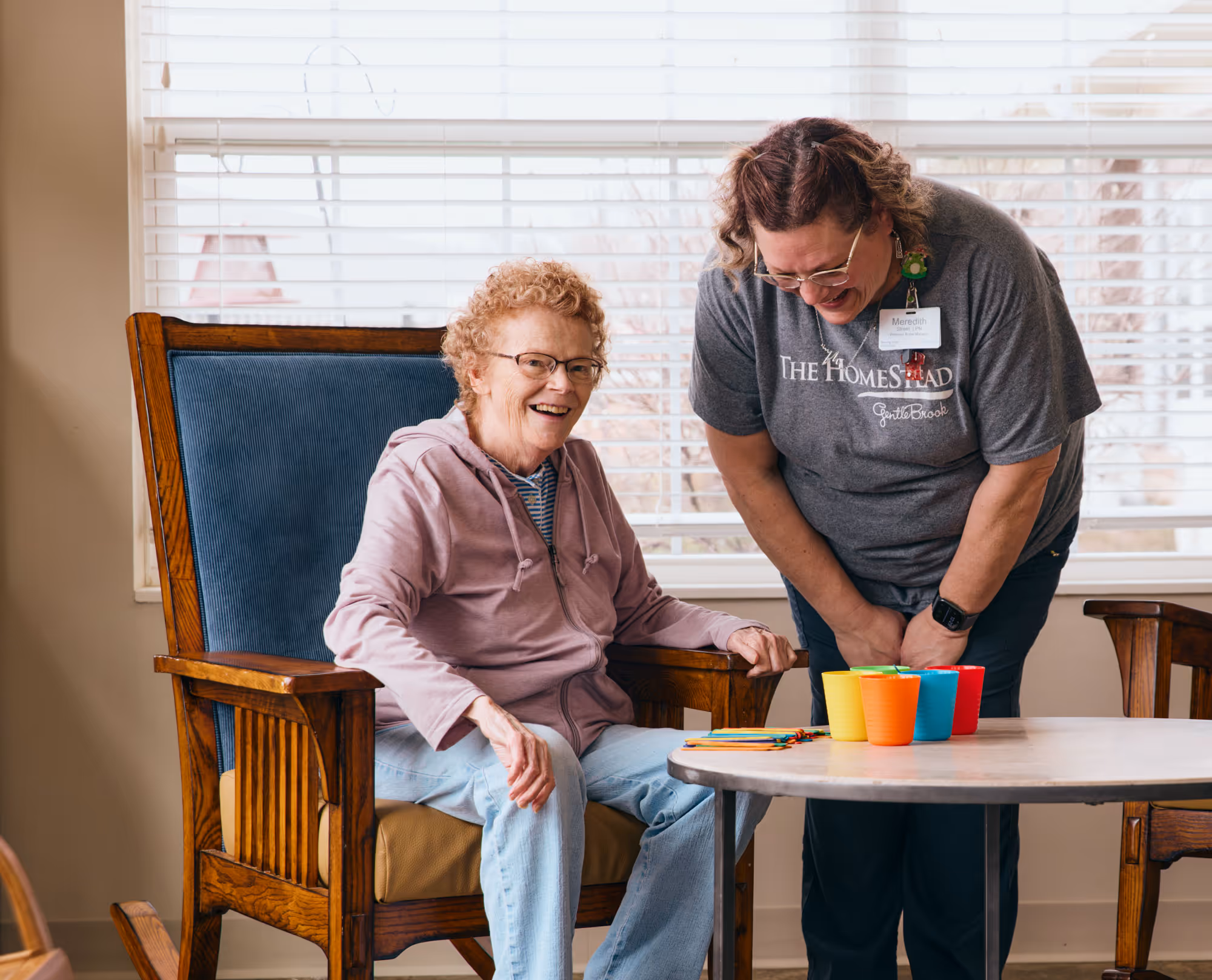 Staff member at The HomeStead at GentleBrook assisting a senior woman seated at a table, engaging in a creative or planting activity in a supportive memory care environment